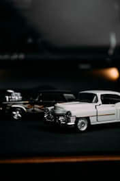 A close-up of two vintage toy cars placed on a dark surface. The focus is on a white classic car in the foreground with chrome detailing and elegant design. In the background, a black car with flames painted on its side is partially visible, adding a contrasting element to the scene.