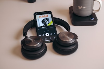 A pair of black over-ear headphones rests on a white surface beside a smartphone displaying an audiobook or podcast with a person's image on the screen. Nearby, there is a grey mug with the word 'offline' placed on a black wireless charging pad.