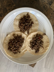Three small round tortillas topped with a serving of cooked minced meat are arranged on a white ceramic plate. The plate rests on a surface with a dark, marbled texture, and the background includes light-colored wooden flooring.