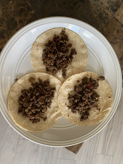 Three small round tortillas topped with a serving of cooked minced meat are arranged on a white ceramic plate. The plate rests on a surface with a dark, marbled texture, and the background includes light-colored wooden flooring.