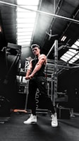 A muscular person in a gym setting is seen flexing their arms. The gym has high ceilings with skylights, allowing natural light to illuminate parts of the room. Workout equipment such as a power rack is visible in the background. The person is wearing a black sleeveless shirt, dark pants, and white high-top sneakers.