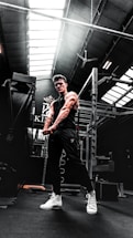 A muscular person in a gym setting is seen flexing their arms. The gym has high ceilings with skylights, allowing natural light to illuminate parts of the room. Workout equipment such as a power rack is visible in the background. The person is wearing a black sleeveless shirt, dark pants, and white high-top sneakers.