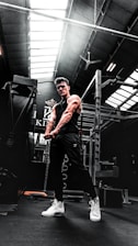 A muscular person in a gym setting is seen flexing their arms. The gym has high ceilings with skylights, allowing natural light to illuminate parts of the room. Workout equipment such as a power rack is visible in the background. The person is wearing a black sleeveless shirt, dark pants, and white high-top sneakers.