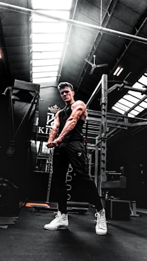 A muscular person in a gym setting is seen flexing their arms. The gym has high ceilings with skylights, allowing natural light to illuminate parts of the room. Workout equipment such as a power rack is visible in the background. The person is wearing a black sleeveless shirt, dark pants, and white high-top sneakers.