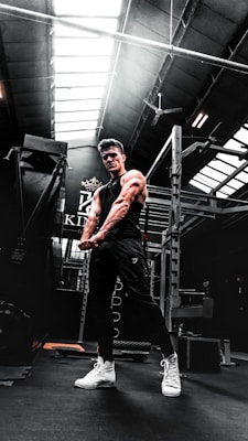 A muscular person in a gym setting is seen flexing their arms. The gym has high ceilings with skylights, allowing natural light to illuminate parts of the room. Workout equipment such as a power rack is visible in the background. The person is wearing a black sleeveless shirt, dark pants, and white high-top sneakers.