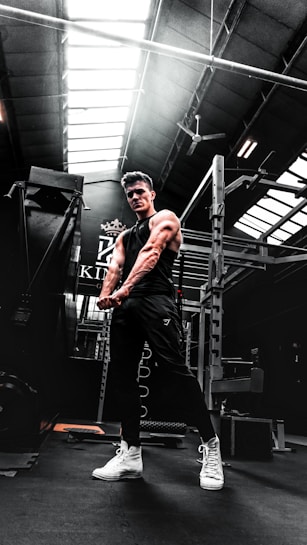 A muscular person in a gym setting is seen flexing their arms. The gym has high ceilings with skylights, allowing natural light to illuminate parts of the room. Workout equipment such as a power rack is visible in the background. The person is wearing a black sleeveless shirt, dark pants, and white high-top sneakers.
