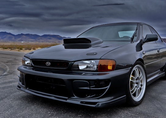 A sleek black sports car positioned on a deserted road with a mountainous landscape in the background. The sky is overcast, creating a dramatic and moody atmosphere. The car features a prominent hood scoop and streamlined design elements.