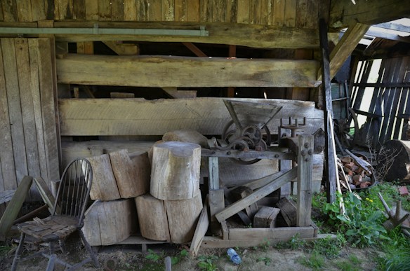 A rustic, weathered wooden structure with stacked logs and a vintage woodcutting machine. An old wooden chair is placed to the side, surrounded by various tools and debris, lending an air of neglect and abandonment. The scene is set in an outdoor environment, with some greenery visible.