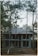 A partially constructed two-story house is situated amidst tall pine trees. The building features an unfinished concrete exterior and a roof with dark shingles. The surrounding area is covered with soil and scattered vegetation, creating a natural, woodland setting.