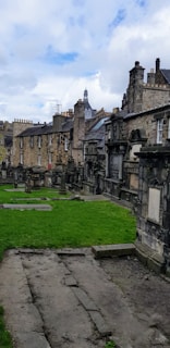 A historic graveyard with old stone tombstones and monuments is surrounded by a row of aged stone buildings in the background. The architecture features classic stonework with chimneys and rooftops visible. The grass is well-kept, and the sky appears partly cloudy with patches of blue and white.