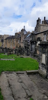 A historic graveyard with old stone tombstones and monuments is surrounded by a row of aged stone buildings in the background. The architecture features classic stonework with chimneys and rooftops visible. The grass is well-kept, and the sky appears partly cloudy with patches of blue and white.