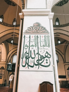 Arabic calligraphy adorns a large vertical panel inside a mosque with intricate floral designs above the script. The building's interior features arched windows and ornamental details.