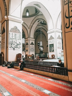 The interior of a grand mosque features large ornate pillars and arches adorned with Arabic calligraphy. People are seated on an expansive red carpet, engaged in reflection or prayer. Soft natural light illuminates the space through arched windows.