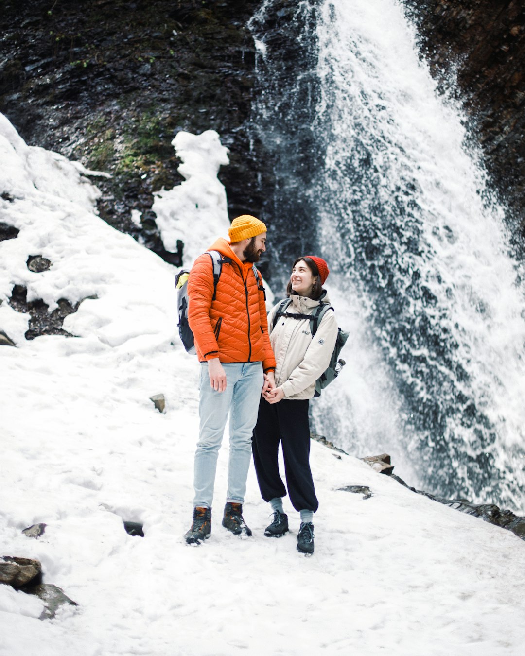 man in orange jacket standing on snow covered ground near waterfalls during daytime