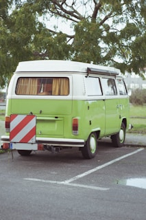 A classic green camper van is parked on a paved area with a leafy tree in the background. The vehicle has a white roof, curtains in the windows, and features a red and white striped warning sign attached to the back.