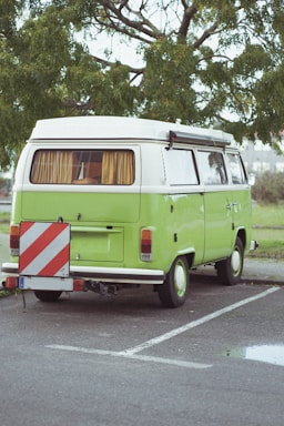 A classic green camper van is parked on a paved area with a leafy tree in the background. The vehicle has a white roof, curtains in the windows, and features a red and white striped warning sign attached to the back.