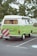 A classic green camper van is parked on a paved area with a leafy tree in the background. The vehicle has a white roof, curtains in the windows, and features a red and white striped warning sign attached to the back.