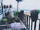 A cozy balcony garden features wooden furniture and a variety of potted plants. The balcony overlooks a cityscape with buildings in the background. Lush green foliage and vibrant pink flowers add a touch of nature to the urban setting.