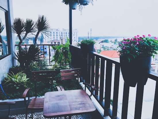 A cozy balcony garden features wooden furniture and a variety of potted plants. The balcony overlooks a cityscape with buildings in the background. Lush green foliage and vibrant pink flowers add a touch of nature to the urban setting.