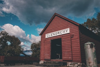 A rustic red wooden building with a sign reading 'Glenorchy' stands prominently against a dramatic sky filled with scattered clouds. The structure has a stone foundation and is surrounded by trees. Off to the side, an old, weathered metal tank adds to the rural, historical ambiance.