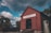 A rustic red wooden building with a sign reading 'Glenorchy' stands prominently against a dramatic sky filled with scattered clouds. The structure has a stone foundation and is surrounded by trees. Off to the side, an old, weathered metal tank adds to the rural, historical ambiance.