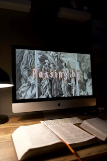 A dimly lit desk setup with an Apple computer displaying an artwork of figures, possibly religious, with the text 'Passing by'. The desk features an open book, lit by a modern desk lamp, alongside a keyboard and another closed book.