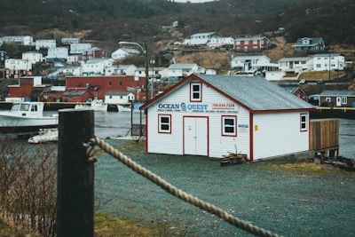 A small coastal town showcases a white building with red trim labeled 'Ocean Quest', situated near the water. Several houses populate the hillside in the background, displaying a mix of white and red exteriors. A lone boat is docked nearby, and the scene is enveloped in a muted, overcast ambiance.