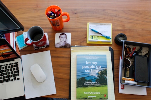 An organized wooden desk features a laptop, a white mouse on a pad, a red coffee mug filled with coffee, a book titled 'Let My People Go Surfing', a tray containing various small items like glasses and pens, a photo coaster with a person's image, a mug with assorted pens, a yellow notepad with notes, and a calendar opened to March 4, 2020.
