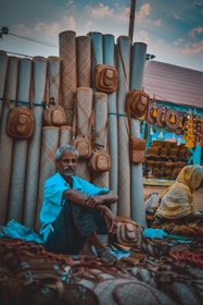 A market scene featuring a vendor sitting among woven goods, including mats and baskets. The vendor appears relaxed, surrounded by an array of handcrafted items that are predominantly made from natural fibers. Another person, dressed in a yellow garment, is partially visible, engaged with the display. The setting appears vibrant and rustic, with a canopy overhead and colorful decorations.