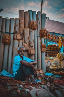 A market scene featuring a vendor sitting among woven goods, including mats and baskets. The vendor appears relaxed, surrounded by an array of handcrafted items that are predominantly made from natural fibers. Another person, dressed in a yellow garment, is partially visible, engaged with the display. The setting appears vibrant and rustic, with a canopy overhead and colorful decorations.