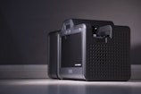 A close-up of a modern electronic device with a sleek and compact design, featuring a black perforated casing and a small display screen on the front. The device is placed on a flat surface against a neutral background, illuminated by soft lighting.