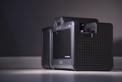 A close-up of a modern electronic device with a sleek and compact design, featuring a black perforated casing and a small display screen on the front. The device is placed on a flat surface against a neutral background, illuminated by soft lighting.