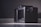 A close-up of a modern electronic device with a sleek and compact design, featuring a black perforated casing and a small display screen on the front. The device is placed on a flat surface against a neutral background, illuminated by soft lighting.