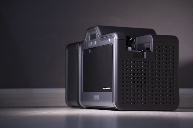 A close-up of a modern electronic device with a sleek and compact design, featuring a black perforated casing and a small display screen on the front. The device is placed on a flat surface against a neutral background, illuminated by soft lighting.