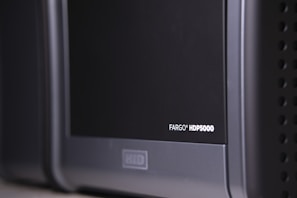 A close-up view of a black and gray electronic device, with the label 'FARGO HDP5000' visible on its surface. The device features a sleek design with rounded edges and some ventilation holes on the side.