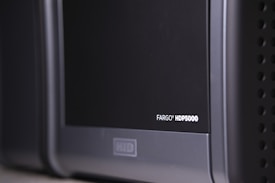 A close-up view of a black and gray electronic device, with the label 'FARGO HDP5000' visible on its surface. The device features a sleek design with rounded edges and some ventilation holes on the side.