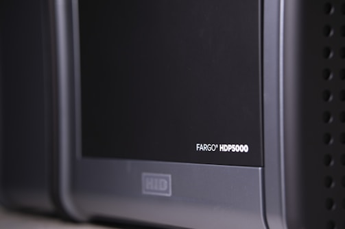 A close-up view of a black and gray electronic device, with the label 'FARGO HDP5000' visible on its surface. The device features a sleek design with rounded edges and some ventilation holes on the side.