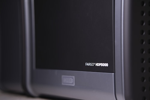 A close-up view of a black and gray electronic device, with the label 'FARGO HDP5000' visible on its surface. The device features a sleek design with rounded edges and some ventilation holes on the side.