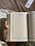 A hand holds an open book with Arabic text on both visible pages, adorned with decorative blue borders. The background features a fabric with a floral pattern, and there is a bookmark with floral design in the upper right corner.