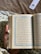 A hand holds an open book with Arabic text on both visible pages, adorned with decorative blue borders. The background features a fabric with a floral pattern, and there is a bookmark with floral design in the upper right corner.