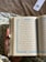 A hand holds an open book with Arabic text on both visible pages, adorned with decorative blue borders. The background features a fabric with a floral pattern, and there is a bookmark with floral design in the upper right corner.