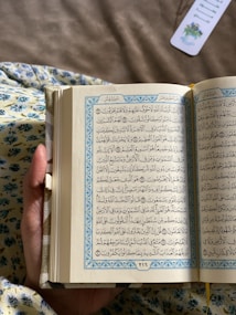 A hand holds an open book with Arabic text on both visible pages, adorned with decorative blue borders. The background features a fabric with a floral pattern, and there is a bookmark with floral design in the upper right corner.