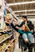 In a grocery store aisle filled with a variety of packaged goods, a young woman sits playfully in a shopping cart reaching for an item on the shelf. A young man stands beside her, also reaching for a product, with a look of amusement. The scene is lively and dynamic, capturing a moment of everyday shopping transformed into a playful experience.