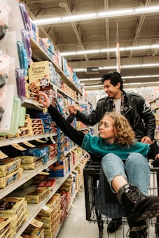 In a grocery store aisle filled with a variety of packaged goods, a young woman sits playfully in a shopping cart reaching for an item on the shelf. A young man stands beside her, also reaching for a product, with a look of amusement. The scene is lively and dynamic, capturing a moment of everyday shopping transformed into a playful experience.