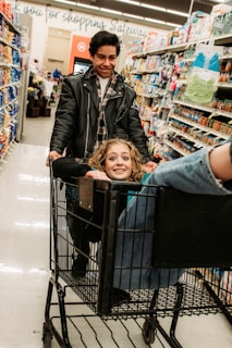A young man pushes a shopping cart with a joyful woman sitting inside. She has curly hair and is smiling up at the camera while he looks down at her, also smiling. They are in a grocery store aisle surrounded by various products on shelves.