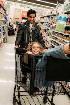 A young man pushes a shopping cart with a joyful woman sitting inside. She has curly hair and is smiling up at the camera while he looks down at her, also smiling. They are in a grocery store aisle surrounded by various products on shelves.