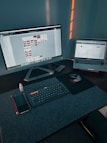 A workspace with a large monitor displaying a design program and a laptop showing a website. On the desk, there is a wireless keyboard, a mouse on a mouse pad, a smartphone, a red pencil, and a small bottle of eye drops. Soft lighting casts shadows on the wall behind the setup.