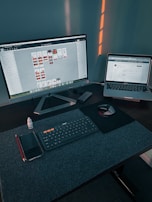A workspace with a large monitor displaying a design program and a laptop showing a website. On the desk, there is a wireless keyboard, a mouse on a mouse pad, a smartphone, a red pencil, and a small bottle of eye drops. Soft lighting casts shadows on the wall behind the setup.