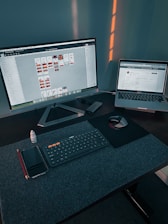 A workspace with a large monitor displaying a design program and a laptop showing a website. On the desk, there is a wireless keyboard, a mouse on a mouse pad, a smartphone, a red pencil, and a small bottle of eye drops. Soft lighting casts shadows on the wall behind the setup.