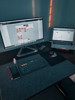 A workspace with a large monitor displaying a design program and a laptop showing a website. On the desk, there is a wireless keyboard, a mouse on a mouse pad, a smartphone, a red pencil, and a small bottle of eye drops. Soft lighting casts shadows on the wall behind the setup.